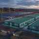 A large Bitcoin mining facility powered by wind turbines, solar panels, and high-voltage transmission lines, with thousands of ASIC mining rigs illustrating how cleaner energy can support expanding Bitcoin mining activity rather than reducing its overall environmental footprint.